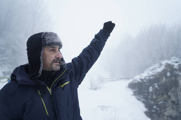 A smiling young man wearing a black winter coat with a fur-lined hood stands outdoors in a snowy, forested setting. The calm expression conveys warmth, resilience, and comfort in cold weather.