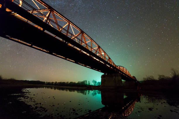 istock Illuminated metal bridge on concrete supports reflected in water on dark starry sky with Milky Way constellation background. Night photography concept. 1132926555