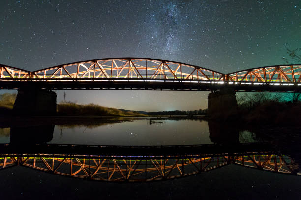 istock Illuminated metal bridge on concrete supports reflected in water on dark starry sky with Milky Way constellation background. Night photography concept. 1132926563