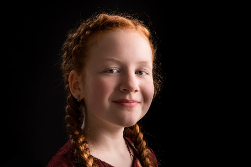 Portrait of smiling girl. Female kid is with redhead braided hair. She is against black background.