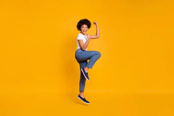 Full length body size side profile photo jumping high beautiful she her lady yelling loud voice legs fists hands arms up win wearing casual jeans denim white t-shirt clothes isolated yellow background.