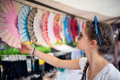 Tourist buying souvenirs on flea market in Mallorca, Spain. Teenage girl is browsing selection of Spanish fans.\nNikon D850