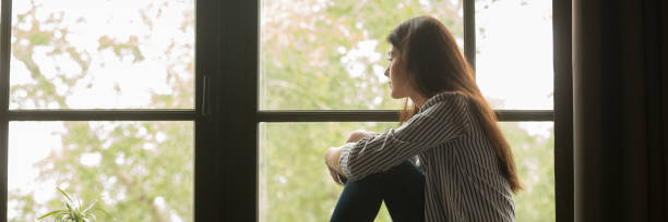 Sad young woman sitting looking out window and thinking Horizontal photo one teen lonely woman sitting at home on sill look at window feels sad depressed frustrated thinking about problem panoramic banner for website header design with copy space for text abortion photos stock pictures, royalty-free photos & images
