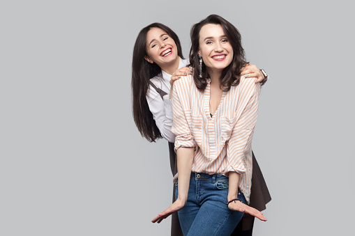 Happy beautiful brunette best friends in casual style standing and having fun together and and looking at camera with toothy smile and good feelings. indoor studio shot, isolated on grey background.