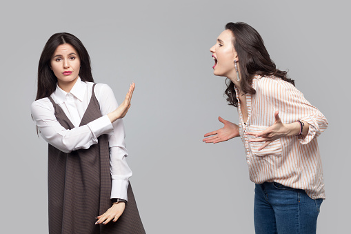 Portrait of young girl looking at camera and showing stop or ignore to another girl who screaming at her. conflict between two friend or partners. indoor studio shot, isolated on grey background