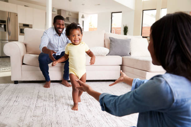 Family At Home Encouraging Baby Daughter To Take First Steps Family At Home Encouraging Baby Daughter To Take First Steps first steps stock pictures, royalty-free photos & images