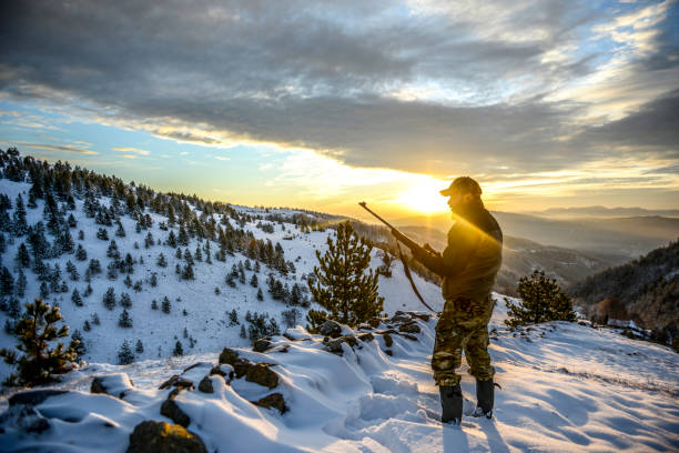 Hunter observing a beautiful vista while standing on a mountain ridge covered with snow Huntsman standing on a snow-capped mountain ridge and observing a wonderful sunset. hunting stock pictures, royalty-free photos & images