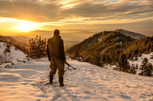 Hunter standing on a snow-capped mountain top observing a serene view at dawn