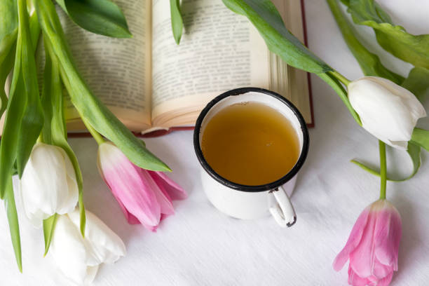 white and pink tulips,  mug of tea, book on white sheets, top view