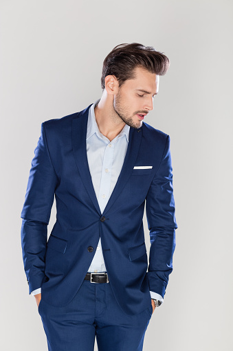 Portrait of handsome young businessman standing with his hands in pocket and looking down against gray background