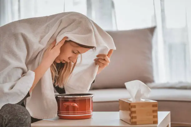 Health care, flu, hygiene and people concept - Photo of sick brown hair woman at home inhaling over a bowl. Rhinitis treatment at home by inhalation. Young woman inhaling chamomile tea steam as an alternative therapy or traditional cure. Young woman inhales steam to treat a blocked nose during the day.