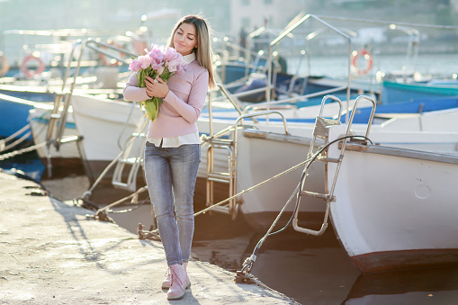 Beautiful girl in spring or autumn, by the sea in the boat Bay. With a bouquet of flowers in his hands and tight stylish jeans and a pink sweater
