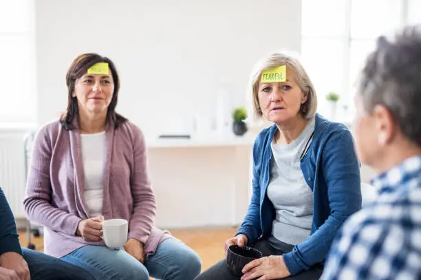 Photo of Men and women sitting in a circle during group therapy, adhesive notes on forehead.