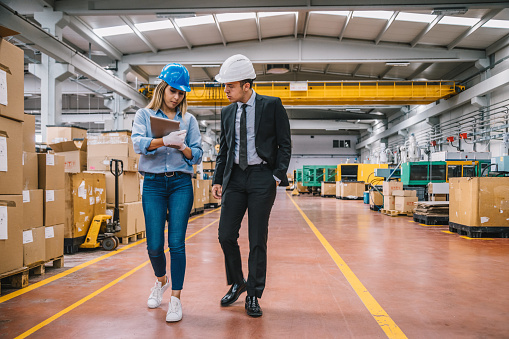 Young warehouse worker supervisor businessman and engineer woman working, walking, talking and looking at digital tablet in a large warehouse in factory