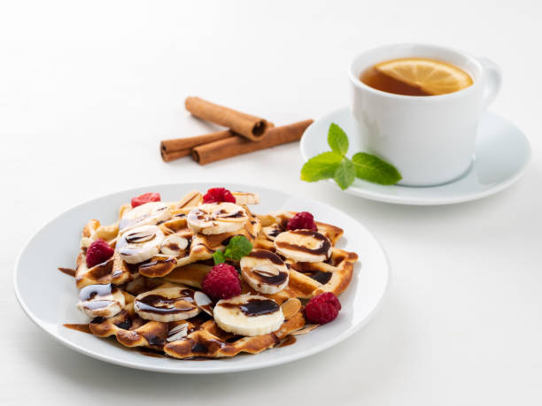 istock Belgian waffles with raspberries, banana,  chocolate syrup.  Breakfast with tea on white background, side view 1132980630