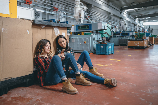 Portrait of young beautiful industrial worker women friends sitting on a floor and having fun with smartphones looking social media contents, discuss about people while looking at phone screen on break time a modern factory plant building. XXXL