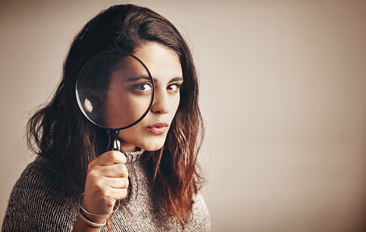 Studio portrait of a young woman looking through a magnifying glass against a brown background