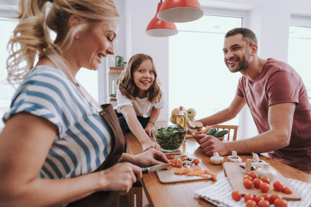 Happy family spending time together and cooking dinner at home Beautiful blond lady in apron cutting carrot with knife while her adorable daughter trying to steal ingredients. Bearded man looking at wife and smiling family dinners and cooking stock pictures, royalty-free photos & images