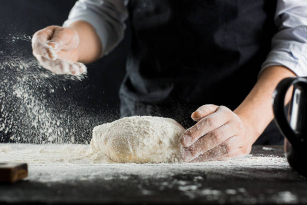 ein koch in einer schwarzen schürze streut mehl auf den küchentisch mit mehl - bread kneading making human hand stock-fotos und bilder