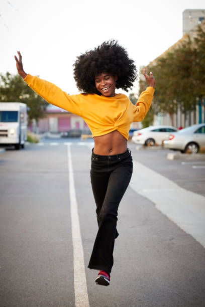 istock Young black woman with afro jumps, looking to camera, while crossing street, front view, vertical 1133003258