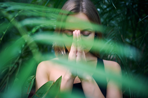Portrait of cute young woman meditating and doing namaste hand in jungle. Sunny day.