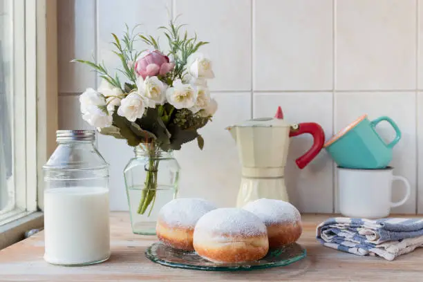 Photo of Three fresh doughnuts on a glass plate with coffee cups, moka pot, flowers and milk bottle in background, by the window
