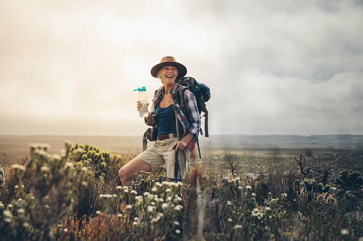 Smiling woman taking a break while hiking in the countryside drinking water. Cheerful senior woman wearing backpack and hat standing on a hill holding a water bottle.
