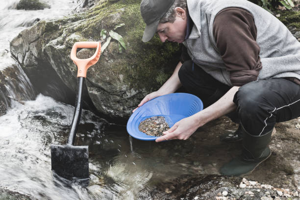 Man with blue pan for gold or batea, rubber boots and shovel, is looking for gold in a creek. Gold panning. Moody instagram look