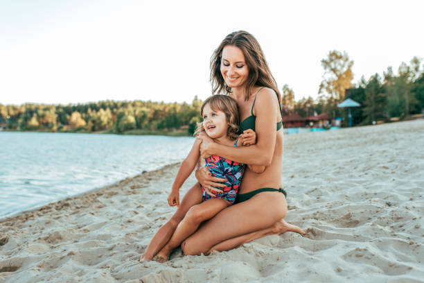 A young mother plays with her daughter girl of 2-5 years, on beach in summer, an outdoor resort by the lake. Happy rest on the weekend. Bright swimsuit on child. Parents love family and parenting. stock photo