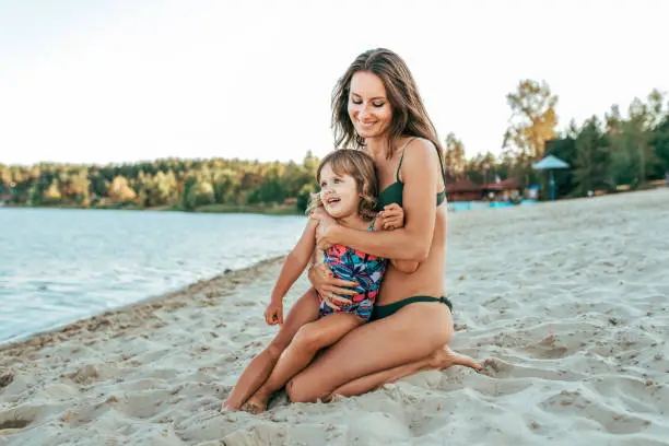 Photo of A young mother plays with her daughter girl of 2-5 years, on beach in summer, an outdoor resort by the lake. Happy rest on the weekend. Bright swimsuit on child. Parents love family and parenting.
