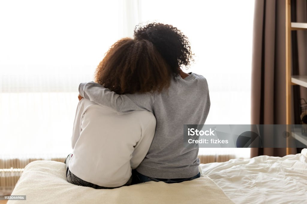 Rear view african mother and daughter embracing sitting on bed Rear back view black mother and daughter embrace sitting on bed at home, older sister consoling younger teen, girl suffers from unrequited love share secrets trustworthy person relative people concept Teenager Stock Photo