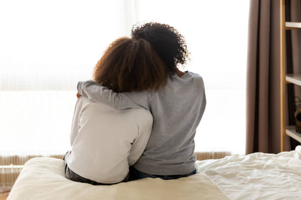 Rear view african mother and daughter embracing sitting on bed Rear back view black mother and daughter embrace sitting on bed at home, older sister consoling younger teen, girl suffers from unrequited love share secrets trustworthy person relative people concept encouragement stock pictures, royalty-free photos & images