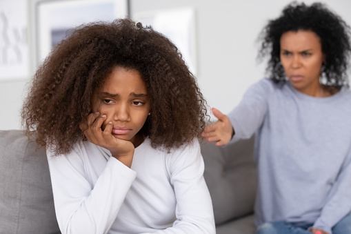 Close up focus african american adolescent girl feels badly melancholic sad and frustrated. On background angry mother accusing scolding reprimanding teen daughter. Teenager behavior problems concept