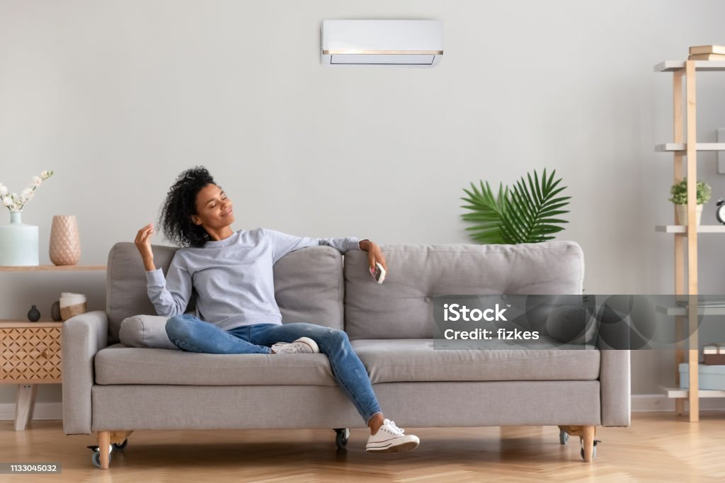 African young relaxed woman sitting on couch breathing fresh air African american relaxed woman sitting on comfortable couch in living room at modern home holds air conditioner remote control enjoying breathing fresh cool air at summer or warm air at winter season Sofa Stock Photo