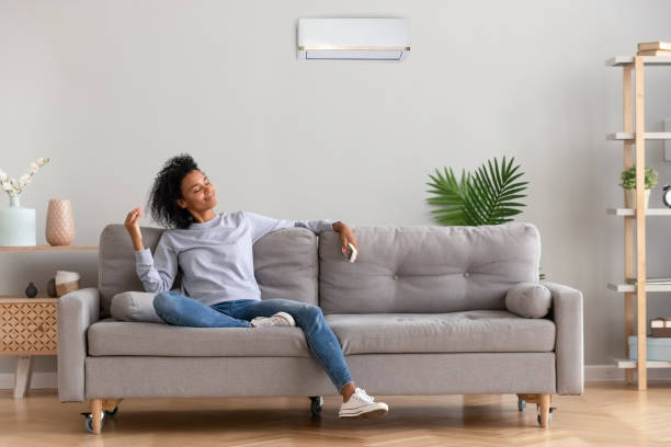 African young relaxed woman sitting on couch breathing fresh air African american relaxed woman sitting on comfortable couch in living room at modern home holds air conditioner remote control enjoying breathing fresh cool air at summer or warm air at winter season remote control photos stock pictures, royalty-free photos & images