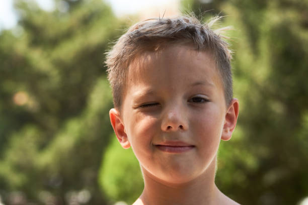 Winking little boy portrait in the park with green trees on background, close-up, outdoors