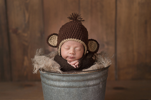 One week old newborn baby boy wearing a brown, crocheted monkey hat. He is sleeping in a galvanized steel bucket and has a slight grin on his face.