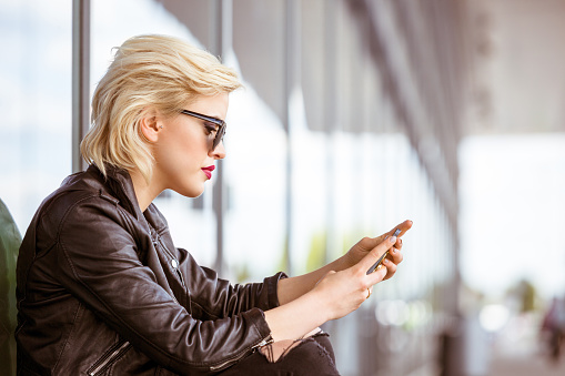 Young woman text messaging on smart phone. Blonde female is sitting at airport. She is wearing leather jacket.