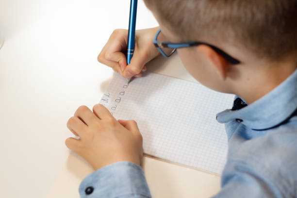 istock Hands of a boy training in writing. Writing letters with a pen in a checkered notebook. 1133074431