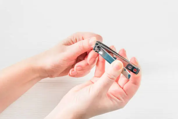 Photo of White Woman's Hand With Healthy Natural Nails Getting Nail Care Procedure. White female hands cutting nails with nail cutter on white wooden background. Beauty Manicure. Personal Hygiene.