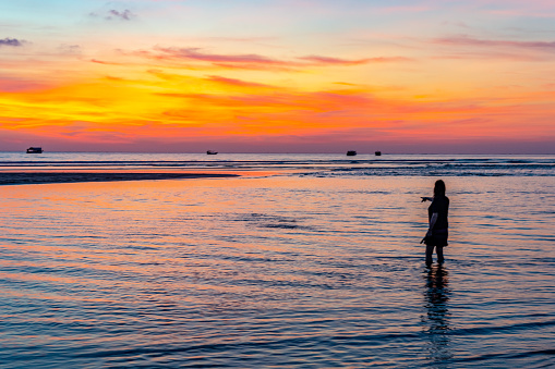 Pakarang beach (Takua Pa) at sunset. A young woman pointing at the stunning sunset whilst wading in the water against a vivid sunset in Thailand, there are long tail boats silhouetted against the night sky in the background.