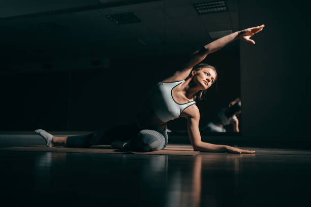fit young caucasian brunette in sportswear sitting on mat in gym and doing side stretching. in background mirror. - posture women side view yoga imagens e fotografias de stock