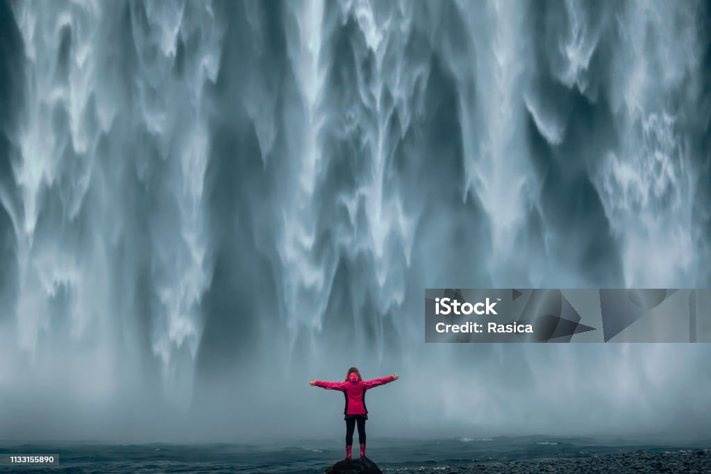 Famous powerful Skogafoss waterfall at south Iceland Iceland landscape photo of brave girl who proudly standing with his arms raised in front of water wall of mighty waterfall. Waterfall Stock Photo