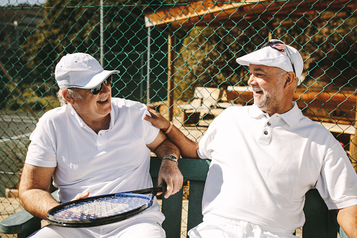 Senior men talking to each other sitting on bench during a game of tennis. Cheerful senior men in tennis clothes taking a break from the game and talking.