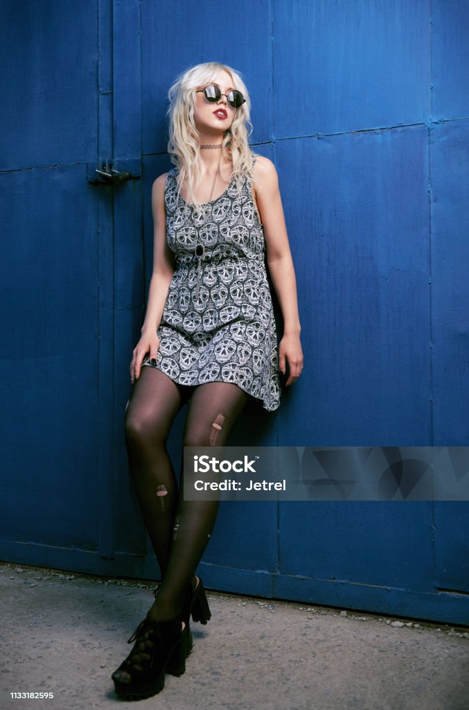 Beautiful young grunge (rock) girl in dress, ripped pantyhose and sunglasses standing at wall. Full length outdoor portrait of informal model Beautiful young grunge (rock) girl in dress, ripped pantyhose and sunglasses standing at wall. Full length outdoor portrait of informal model. Film grain added (not camera noise) Alternative Rock Stock Photo