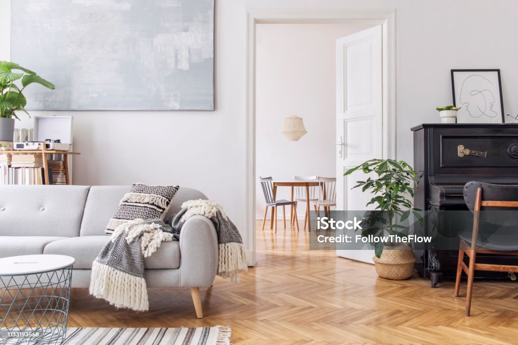 Modern scandianvian decor of living room with design sofa with elegant blanket, coffee table and bookstand on the white wall. Brown wooden parquet. Concept of minimalistic interior with piano. Mock up Modern scandianvian decor of living room. Concept of minimalistic interior. Living Room Stock Photo