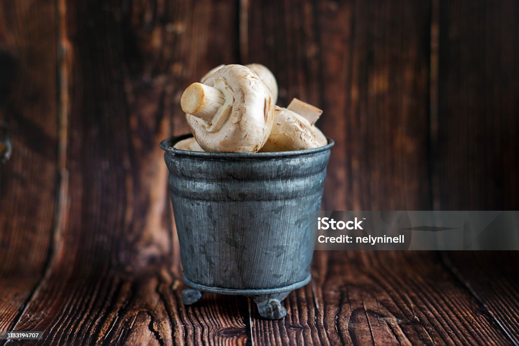 Fresh whole white mushrooms, in a metal bucket with legs on a wooden background, ready for cleaning and cooking for lunch. Fresh whole white mushrooms, in a metal bucket with legs on a wooden background, ready for cleaning and cooking for lunch Agaricus Stock Photo