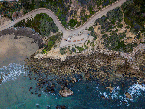 Aerial birds eye view of a rock formation along the southern California coast. There is a walking path down the hillside of the coast. Clear blue and green water with waves crashing against the side of the rock.