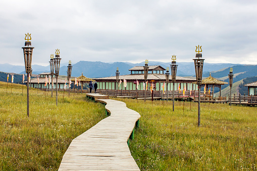Wooden Boardwalk Across Grassland Above Lawn Leads A Couple Of People To Contemporary Mongolian And Tibetan Pavilions.