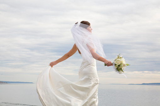 Beautiful, happy, smiling bride in white wedding gown and veil holding a bouquet with water and sky background